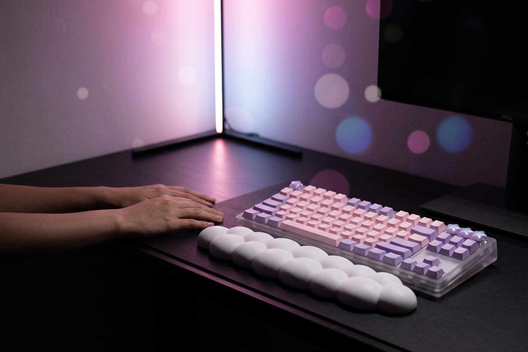 A desk at night lit only by a corner floor lamp glowing soft purple, keyboard and cloud wrist rest visible